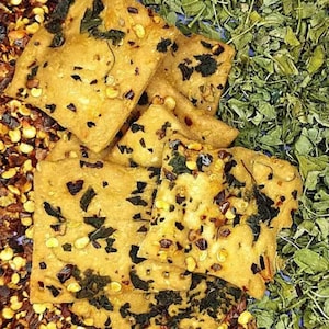 May include: Close-up of square crackers with herbs and spices. The crackers are golden brown with dark green herbs and red pepper flakes. Dried green herbs and red pepper flakes are scattered around the crackers.