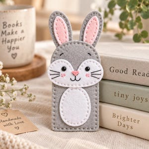 Puede incluir: Un marcapáginas de fieltro gris con forma de conejo, orejas largas, interior de orejas rosa, hocico blanco y vientre blanco. El conejo tiene ojos y bigotes bordados en negro y una nariz rosa. El marcapáginas es rectangular con bordes cosidos.