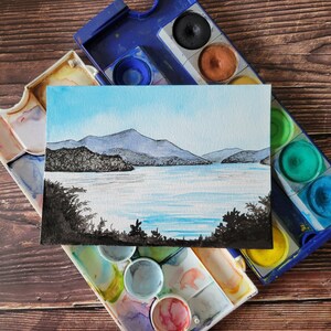 May include: A watercolor painting of a mountain range with a lake in the foreground. The painting is on a white card and is surrounded by a blue and white watercolor palette.
