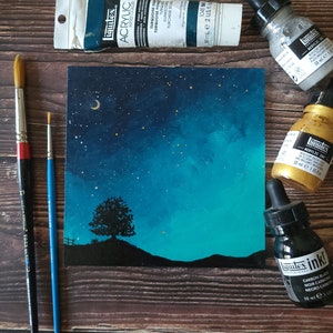 May include: A painting of a night sky with a crescent moon and stars. A single tree is silhouetted in the foreground. The painting is on a canvas and is surrounded by paint tubes and bottles.