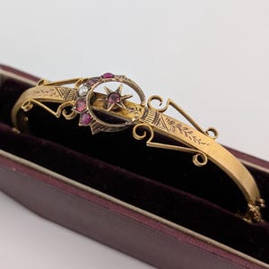 May include: A gold hinged bangle bracelet featuring a crescent moon and star motif. The crescent is set with small red stones and a single white pearl, while the star contains a central red stone. The band has engraved leaf and geometric details.