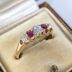 Puede incluir: Un anillo de oro con cinco piedras preciosas: dos claras, dos rojas y una clara. El anillo está engastado en una banda de oro y se muestra en un joyero vintage con un interior color crema.