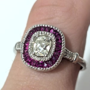 May include: A silver ring featuring a central, square-cut, clear gemstone surrounded by a halo of small, vibrant, magenta-colored gemstones. The ring's band has delicate detailing.