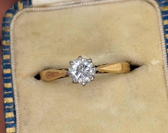 May include: A gold ring featuring a single round brilliant-cut clear gemstone set in a six-prong white metal head. The band has a tapered design with textured, geometric gold shoulders that connect to the central stone setting.