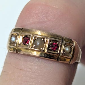 May include: A gold ring with a rectangular design, featuring alternating gemstones. The ring has small, square settings holding red and pearl-coloured stones, with a larger pearl-coloured stone in the centre. The ring is on a finger.
