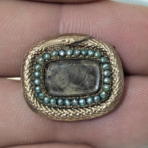 May include: A gold-toned antique brooch with a rectangular center containing a dark, textured material, possibly hair. The rectangle is surrounded by small, teal-colored beads. The brooch has a snake design around the edge.