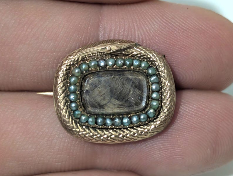 May include: A gold-toned antique brooch with a rectangular center containing a dark, textured material, possibly hair. The rectangle is surrounded by small, teal-colored beads. The brooch has a snake design around the edge.
