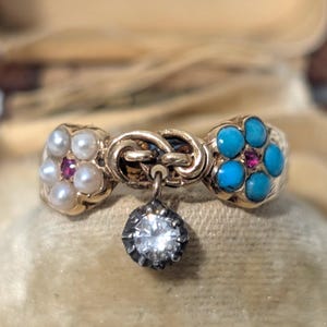 May include: A gold ring with three sections. One section features pearls and a red stone, another has turquoise stones and a red stone, and the center has a diamond pendant. The ring is displayed in a velvet-lined box.