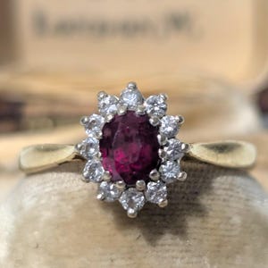 Puede incluir: Un anillo de oro con una gema central de rubí ovalada rodeada por un halo de pequeños diamantes brillantes. El anillo se exhibe sobre un fondo de terciopelo beige suave, resaltando su elegante diseño.