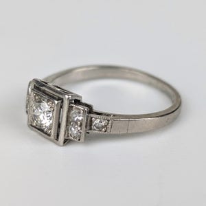 K&ouml;nnte beinhalten: Ein silberner Ring mit einem quadratischen Mittelstein und kleineren Akzentsteinen. Der Ring hat ein klassisches Design mit einem d&uuml;nnen Band und ist vor einem wei&szlig;en Hintergrund platziert. Die Fassung scheint aus Platin oder Wei&szlig;gold zu sein.