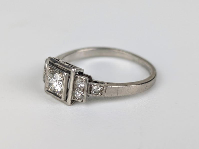 K&ouml;nnte beinhalten: Ein silberner Ring mit einem quadratischen Mittelstein und kleineren Akzentsteinen. Der Ring hat ein klassisches Design mit einem d&uuml;nnen Band und ist vor einem wei&szlig;en Hintergrund platziert. Die Fassung scheint aus Platin oder Wei&szlig;gold zu sein.