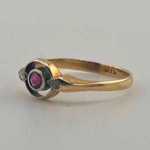 May include: A gold ring featuring a central circular setting with a vibrant pink gemstone. The setting is accented by two small, clear stones on either side, integrated into a smooth, polished gold band with a tapered design.