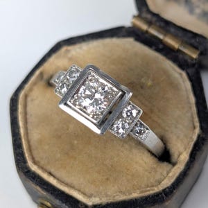 K&ouml;nnte beinhalten: Ein silberner Ring mit einem quadratischen, diamantbesetzten Mittelstein und kleineren Diamanten an den Seiten. Der Ring befindet sich in einer schwarzen Vintage-Schmuckschatulle mit beigem Innenraum. Ein klassisches Schmuckst&uuml;ck.