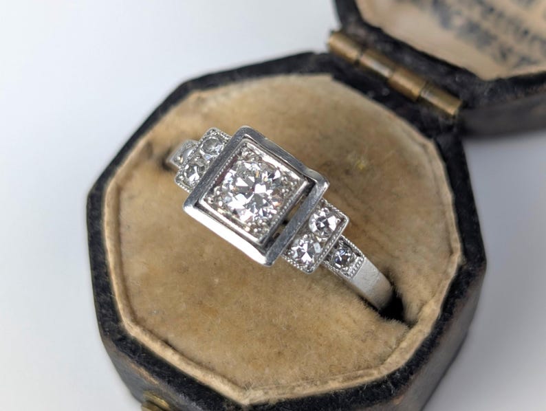 K&ouml;nnte beinhalten: Ein silberner Ring mit einem quadratischen, diamantbesetzten Mittelstein und kleineren Diamanten an den Seiten. Der Ring befindet sich in einer schwarzen Vintage-Schmuckschatulle mit beigem Innenraum. Ein klassisches Schmuckst&uuml;ck.