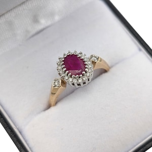 May include: A gold ring featuring a large oval ruby gemstone, encircled by a halo of small diamonds. The ring is set in a gold band with diamond accents. Presented in a black jewellery box with a white satin lining.