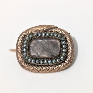 May include: Antique gold-toned brooch with a snake design. The brooch features a rectangular center stone surrounded by small, gray-blue pearls. The snake's body forms an oval shape around the center, with a pin on the back.