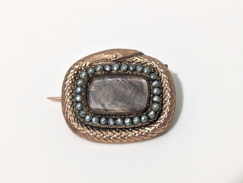 May include: Antique gold-toned brooch with a snake design. The brooch features a rectangular center stone surrounded by small, gray-blue pearls. The snake's body forms an oval shape around the center, with a pin on the back.