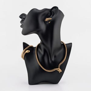 Puede incluir: Un conjunto de joyería en tono dorado que incluye un collar tipo gargantilla con forma de serpiente, un brazalete a juego con forma de serpiente y un pendiente de botón con forma de serpiente. Cada pieza tiene una textura escamosa.