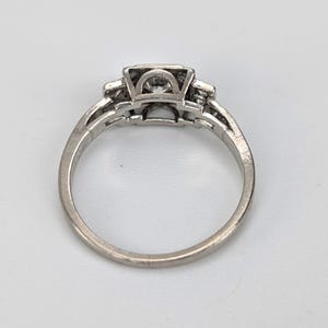 K&ouml;nnte beinhalten: Ein silberner Ring mit einem quadratischen Mittelstein und kleineren Steinen auf beiden Seiten. Der Ring hat ein detailliertes, durchbrochenes Design am Band. Der Ring ist vor einem wei&szlig;en Hintergrund platziert.