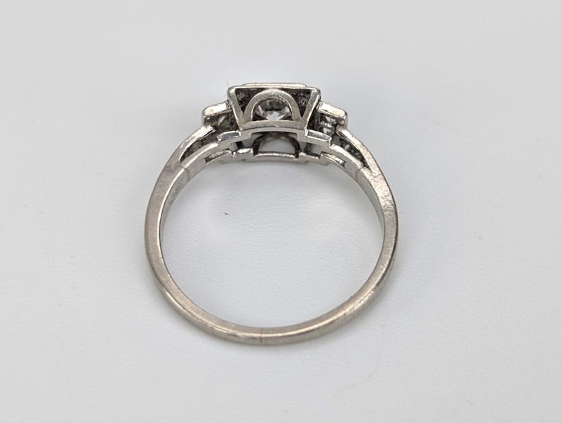 K&ouml;nnte beinhalten: Ein silberner Ring mit einem quadratischen Mittelstein und kleineren Steinen auf beiden Seiten. Der Ring hat ein detailliertes, durchbrochenes Design am Band. Der Ring ist vor einem wei&szlig;en Hintergrund platziert.