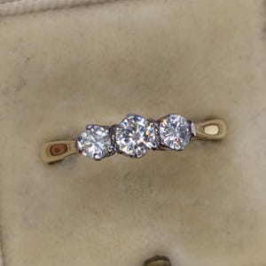 May include: A gold ring featuring three round, clear, faceted stones set in a row. The centre stone is slightly larger than the two flanking stones, and each is held in place by a metal setting. The ring band is smooth and polished gold.