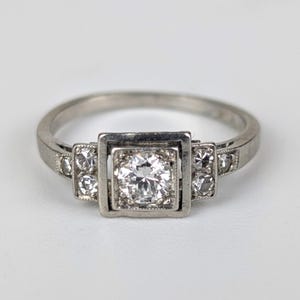 K&ouml;nnte beinhalten: Ein silberner Ring mit quadratischer Mitte, der einen gro&szlig;en, runden, klaren Edelstein enth&auml;lt. Kleinere klare Edelsteine sind auf beiden Seiten des Mittelsteins gesetzt. Der Ring ist vor einem wei&szlig;en Hintergrund platziert.