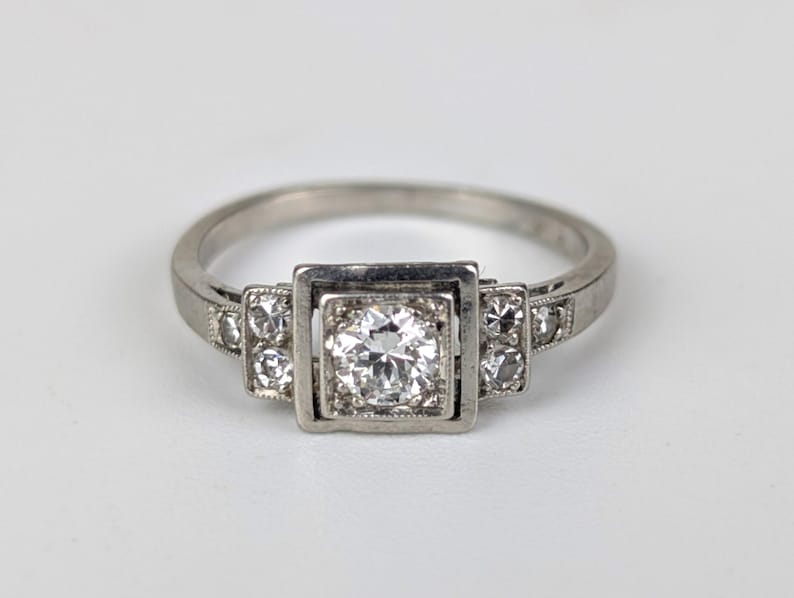 K&ouml;nnte beinhalten: Ein silberner Ring mit quadratischer Mitte, der einen gro&szlig;en, runden, klaren Edelstein enth&auml;lt. Kleinere klare Edelsteine sind auf beiden Seiten des Mittelsteins gesetzt. Der Ring ist vor einem wei&szlig;en Hintergrund platziert.