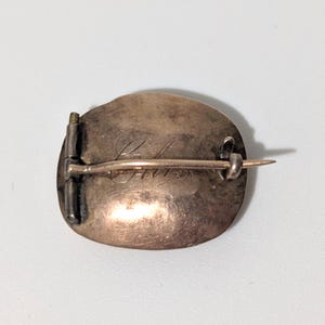 May include: A vintage oval-shaped brooch with a pin closure. The brooch has a tarnished, bronze-colored surface with engraved script. The pin and clasp are silver-toned. The brooch is likely made of metal.