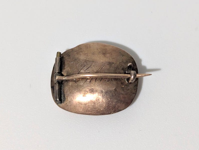 May include: A vintage oval-shaped brooch with a pin closure. The brooch has a tarnished, bronze-colored surface with engraved script. The pin and clasp are silver-toned. The brooch is likely made of metal.