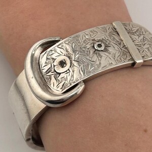 Puede incluir: Una pulsera de metal plateado con diseño de cinturón, que presenta una hebilla pulida y una superficie texturizada con un patrón de hojas. La pulsera incluye una trabilla metálica fija y un detalle circular central.