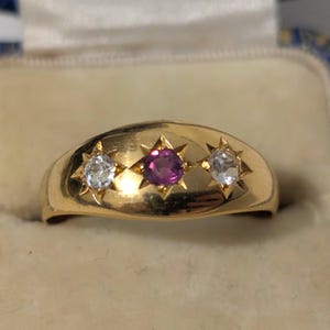 May include: A gold ring featuring a central round-cut pink gemstone flanked by two clear, round-cut white gemstones. Each stone is set within a star-shaped engraving on the smooth, polished gold band, creating a cohesive and detailed design.