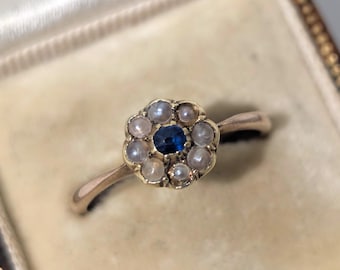 Puede incluir: Un anillo de oro con un diseño floral. La piedra central es de color azul oscuro, rodeada de pequeñas piedras de color claro. El anillo está engastado en una banda de oro, con un engaste en forma de flor.