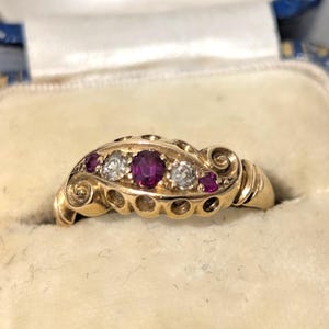 Puede incluir: Un anillo de oro con una piedra preciosa central de color rojo intenso, flanqueada por dos piedras blancas transparentes y dos piedras rojas más pequeñas. La banda presenta un diseño metálico curvo con recortes circulares laterales.