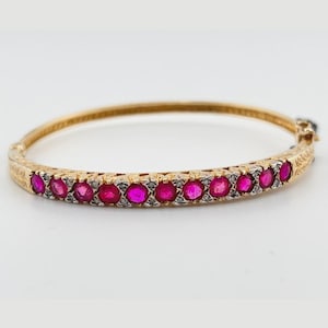 May include: A gold hinged bangle bracelet featuring a row of round, vibrant pink rubies set in silver-toned metal. The gold band displays intricate engraved detailing along the sides and includes a secure clasp mechanism on the right side.