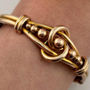 Pode incluir: Uma pulseira de metal em tom dourado com um design de nó central. A pulseira é construída com elementos de fio liso e polido, e inclui pequenos detalhes de contas esféricas integradas na estrutura nas laterais do nó central.