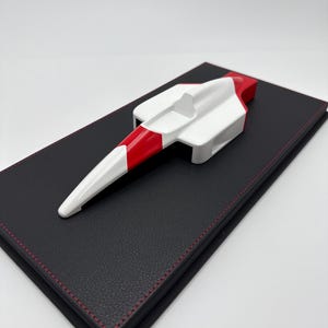 Può includere: Un modello di auto da corsa bianco e rosso è posizionato su un libro nero rilegato in pelle con cuciture rosse. L'auto ha un design aerodinamico, con un muso appuntito e un abitacolo. Il libro fornisce uno sfondo contrastante.