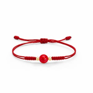 Op de afbeelding: Rode draadarmband met een centrale rode kraal en twee kleine gouden kralen. De armband is verstelbaar met een schuifknoop. Het ontwerp is eenvoudig en elegant.