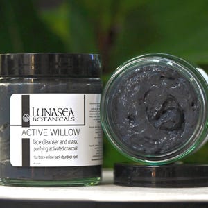 May include: Two jars of Active Willow face cleanser and mask by Lunasea Botanicals. One jar is upright, the other is open and lying on its side, revealing a dark gray, charcoal-based product. The label lists ingredients like tea tree and willow bark.