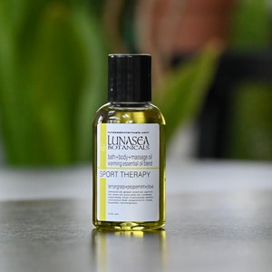 May include: A small, clear glass bottle with a black cap, containing a yellow liquid. The bottle is labeled "LUNASEA BOTANICALS" and "SPORT THERAPY" with the text "lemongrass+peppermint+dove".