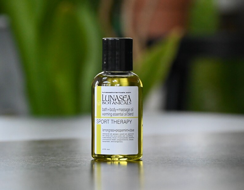 May include: A small, clear glass bottle with a black cap, containing a yellow liquid. The bottle is labeled "LUNASEA BOTANICALS" and "SPORT THERAPY" with the text "lemongrass+peppermint+dove".