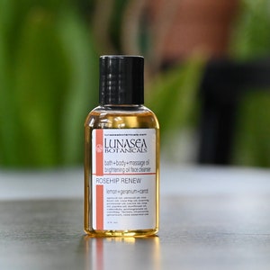 May include: A small amber glass bottle with a black cap and a white label. The label reads "LUNASEA BOTANICALS bath+body+massage oil brightening oil face cleanser ROSEHIP RENEW lemon+geranium+carrot"