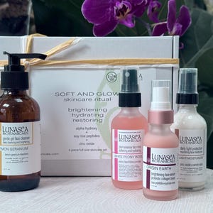 Puede incluir: Un set de cuidado de la piel con una botella marrón con bomba, tres botellas de spray y una caja atada con una cinta. El set incluye productos con ingredientes como geranio limón, peonía blanca y óxido de zinc. La caja dice "SOFT AND GLOW skincare ritual".