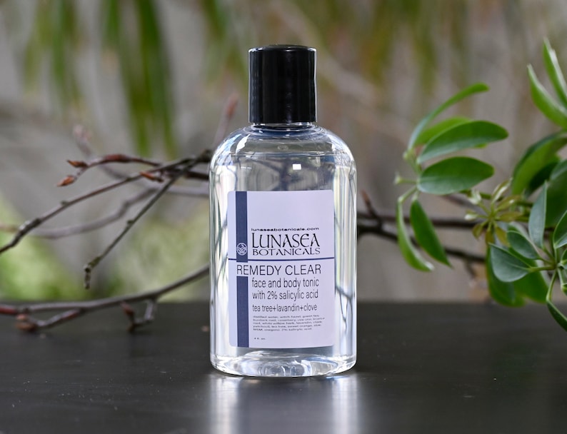 May include: A clear glass bottle with a black cap, labeled "LUNASEA BOTANICALS REMEDY CLEAR face and body tonic with 2% salicylic acid tea tree+lavandin+clove".