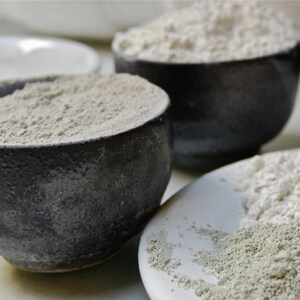 May include: Three bowls filled with different types of white powder. The bowls are made of dark gray ceramic and have a rough texture. The powder in the bowls is a light gray color and has a fine texture.