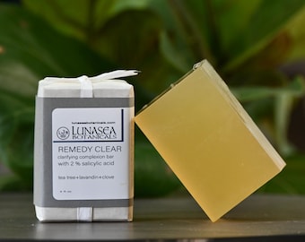 May include: A rectangular bar of translucent yellow soap next to its packaging. The white paper wrapper features a gray label with the text Lunasea Botanicals, Remedy Clear, clarifying complexion bar with 2% salicylic acid, tea tree, lavandin, clove.