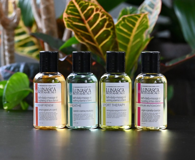 May include: Four amber glass bottles with black lids and white labels. The labels have the text "LUNASEA BOTANICALS" and list different bath and body oil blends: "ROSEHIP RENEW", "BREATHE", "SPORT THERAPY", and "ESERUM ANTIOXIDANT".