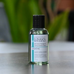 May include: A clear glass bottle with a black cap and a white label. The label reads "LUNASEA BOTANICALS bath+body+massage oil soothing essential oil blend BREATHE lavandin+rosemary+eucalypta".