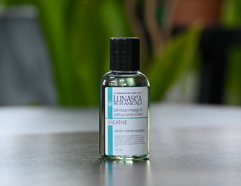 May include: A clear glass bottle with a black cap and a white label. The label reads "LUNASEA BOTANICALS bath+body+massage oil soothing essential oil blend BREATHE lavandin+rosemary+eucalypta".