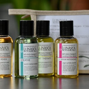 May include: Four small amber glass bottles with black caps and labels. The bottles contain bath and body oils with different scents and benefits. The labels read "LUNASEA BOTANICALS" and list the scent and benefits of each oil. The bottles are arranged in a row on a wooden surface.