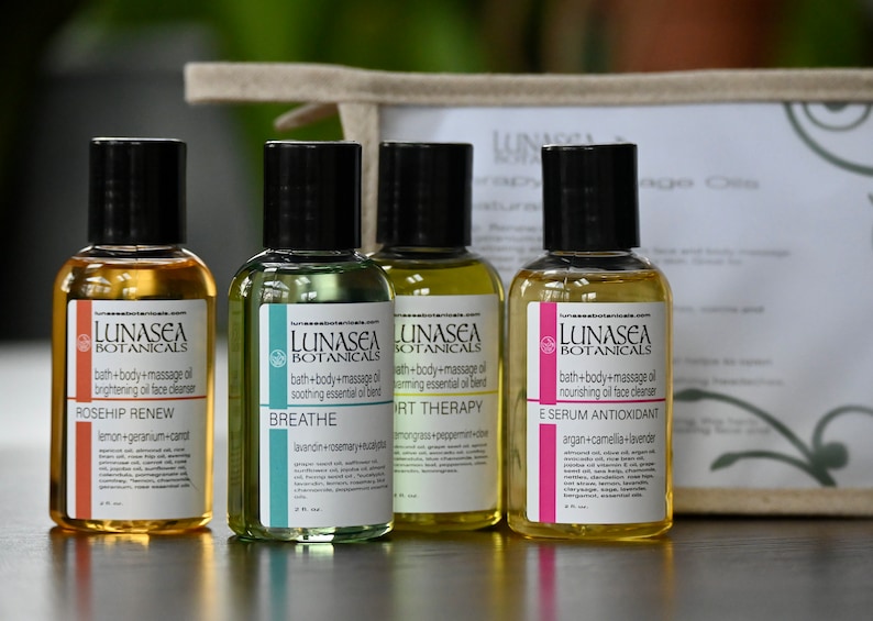 May include: Four small amber glass bottles with black caps and labels. The bottles contain bath and body oils with different scents and benefits. The labels read "LUNASEA BOTANICALS" and list the scent and benefits of each oil. The bottles are arranged in a row on a wooden surface.